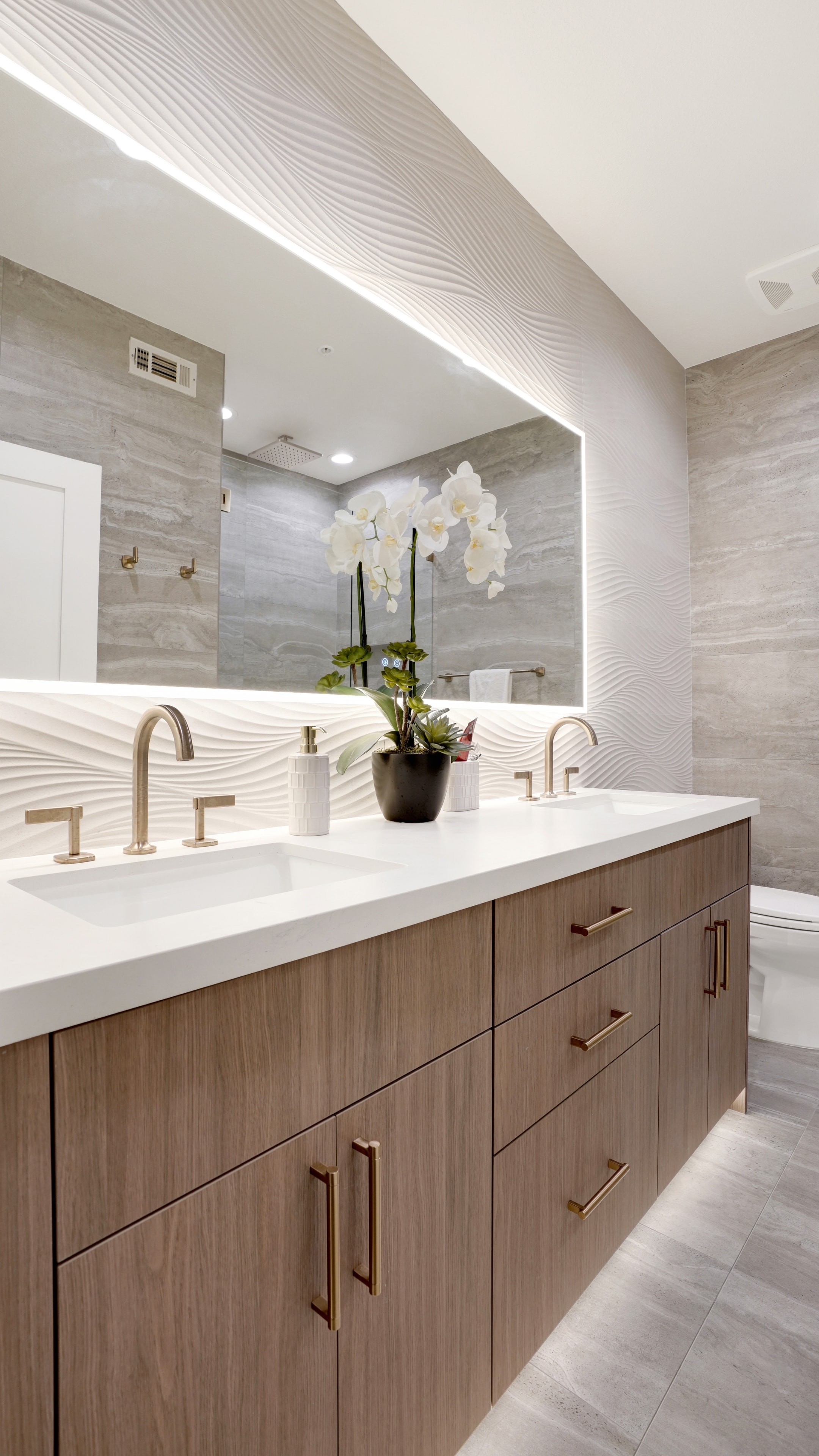 Bathroom remodel in Santa Ana CA — double vanity with wood cabinetry, white quartz countertop, 3D wave tile backsplash, dual brass widespread faucets, and large backlit LED mirror by Main Street Kitchen & Flooring