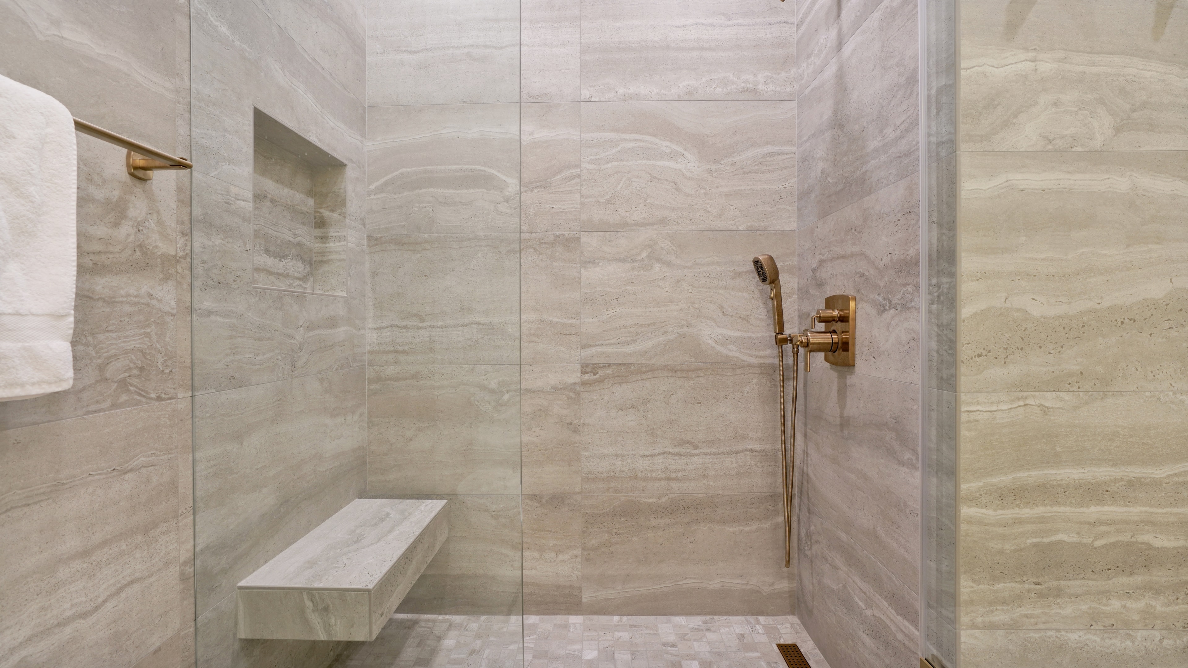 Bathroom remodel in Santa Ana CA — open walk-in shower with travertine slab walls, floating marble bench, brass handheld shower, niche shelf, and mosaic tile floor by Main Street Kitchen & Flooring