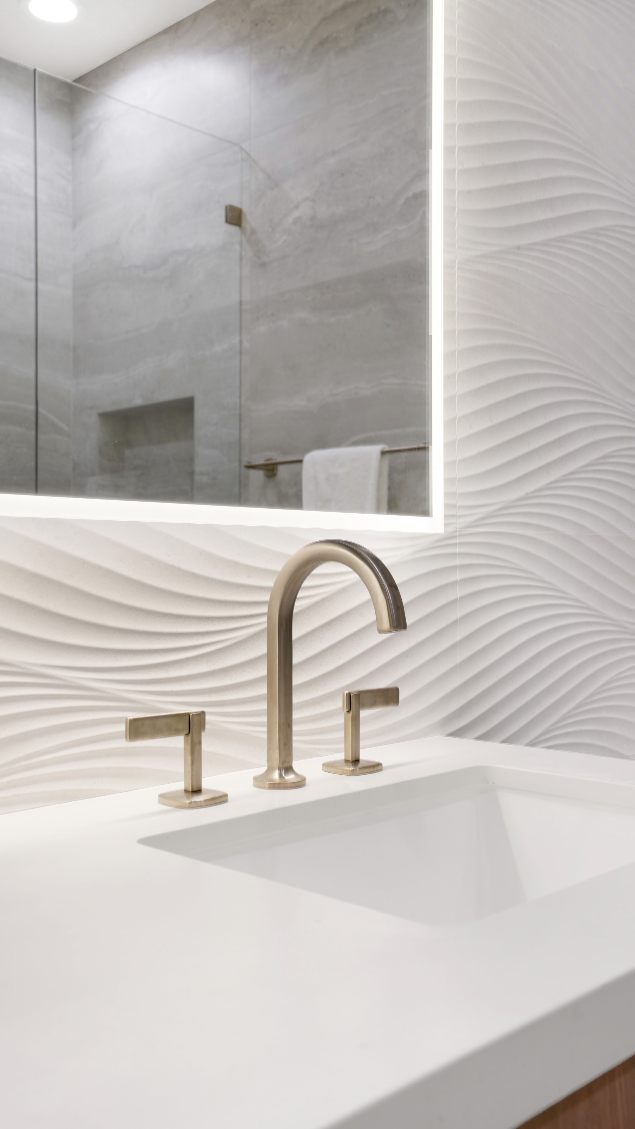Bathroom remodel in Santa Ana CA — closeup of 3D wave texture backsplash tile with brushed brass widespread faucet, undermount sink, and backlit LED mirror reflection by Main Street Kitchen & Flooring