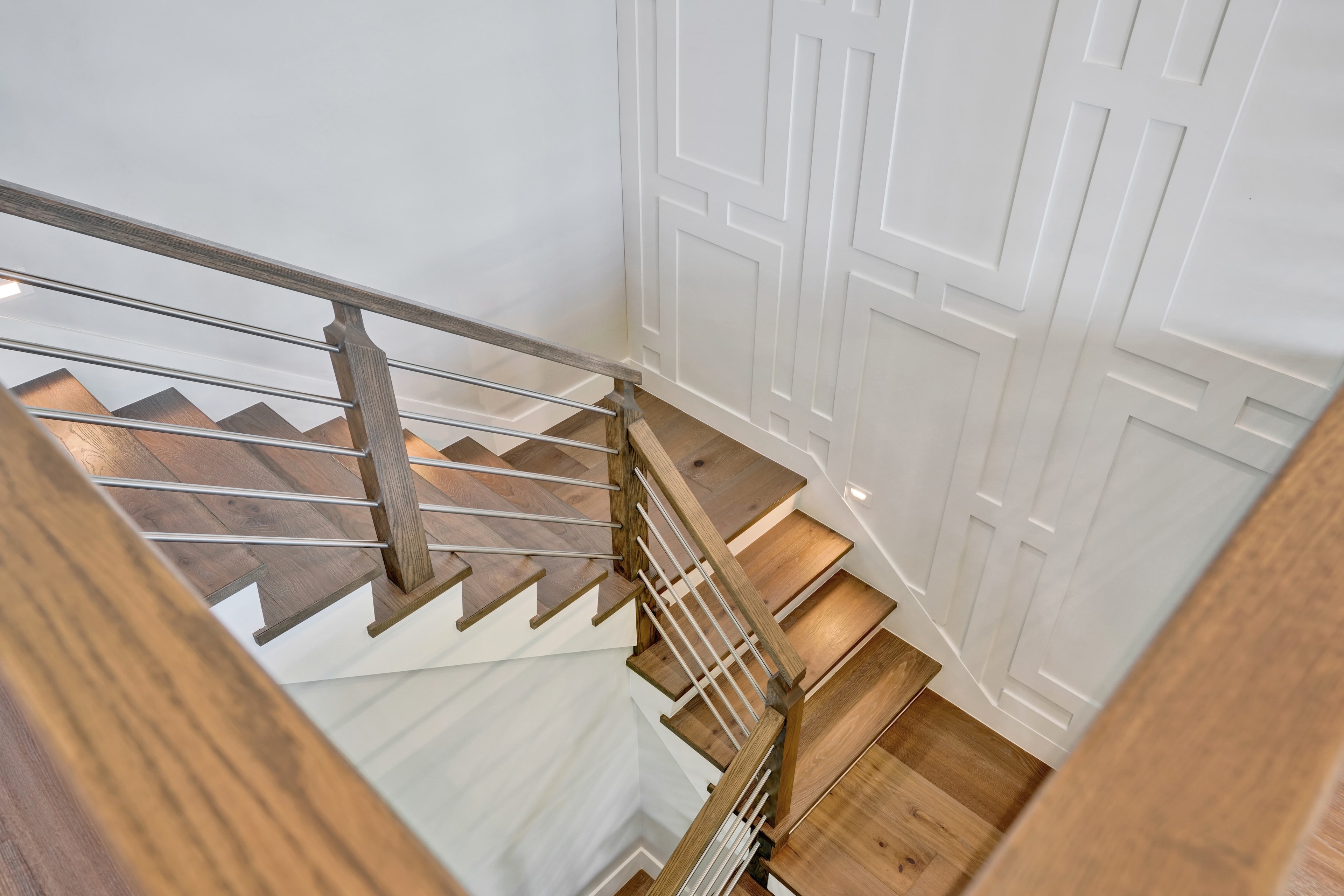 Home remodel in Orange County CA — overhead view of custom staircase with wide-plank wood treads, stained wood newel posts, horizontal steel cable railing, and geometric raised-panel wainscoting accent wall by Main Street Kitchen & Flooring