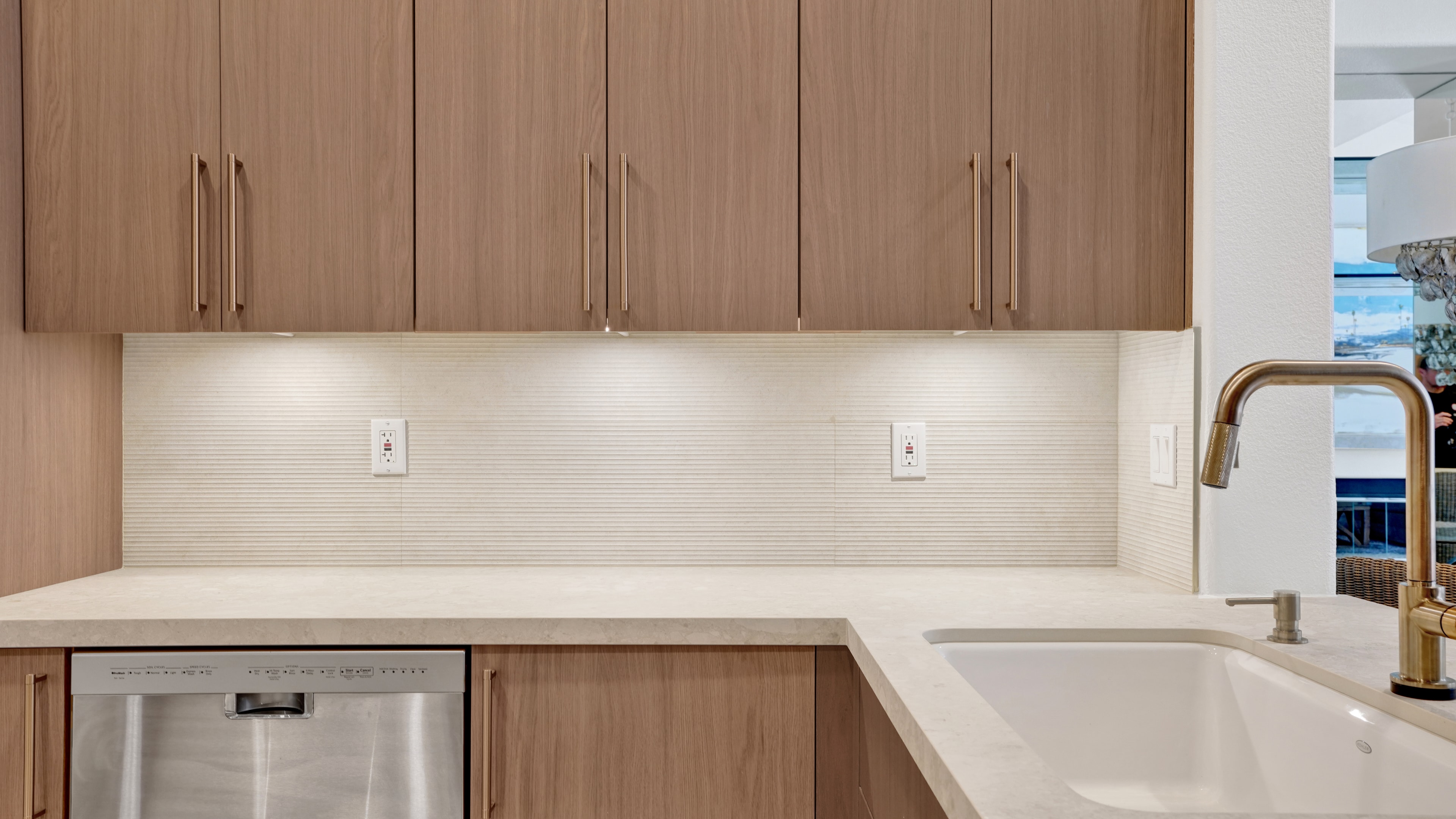 Kitchen remodel in Irvine CA — wood flat-panel upper cabinets with brass pulls, fluted ceramic tile backsplash, white quartz countertop, undermount sink with brass faucet, stainless dishwasher, and ocean view through window by Main Street Kitchen & Flooring