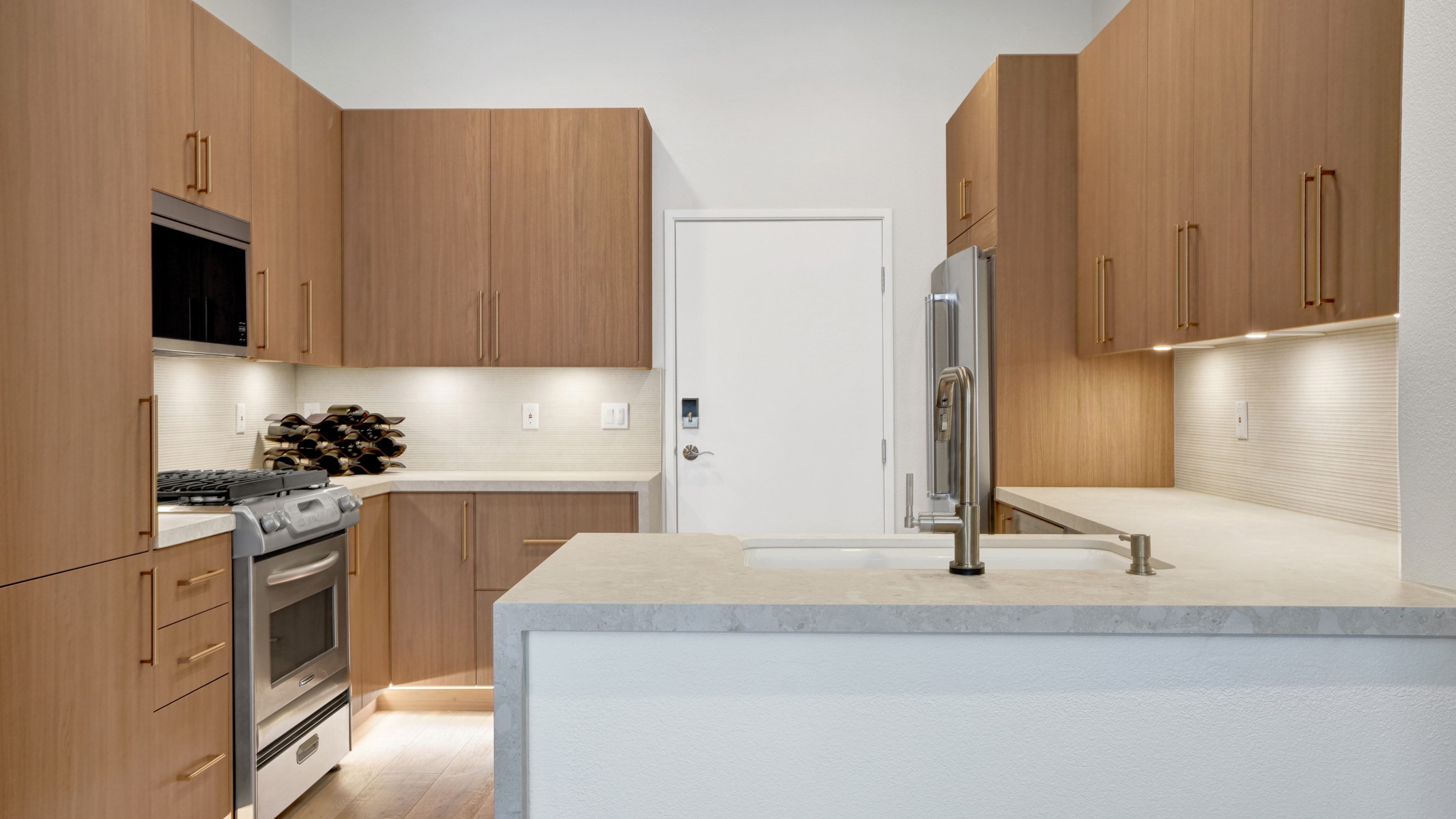 Kitchen remodel in Irvine CA — warm wood flat-panel cabinetry with brass bar pulls, white quartz island countertop, undermount sink with brass faucet, fluted tile backsplash, and stainless steel appliances by Main Street Kitchen & Flooring