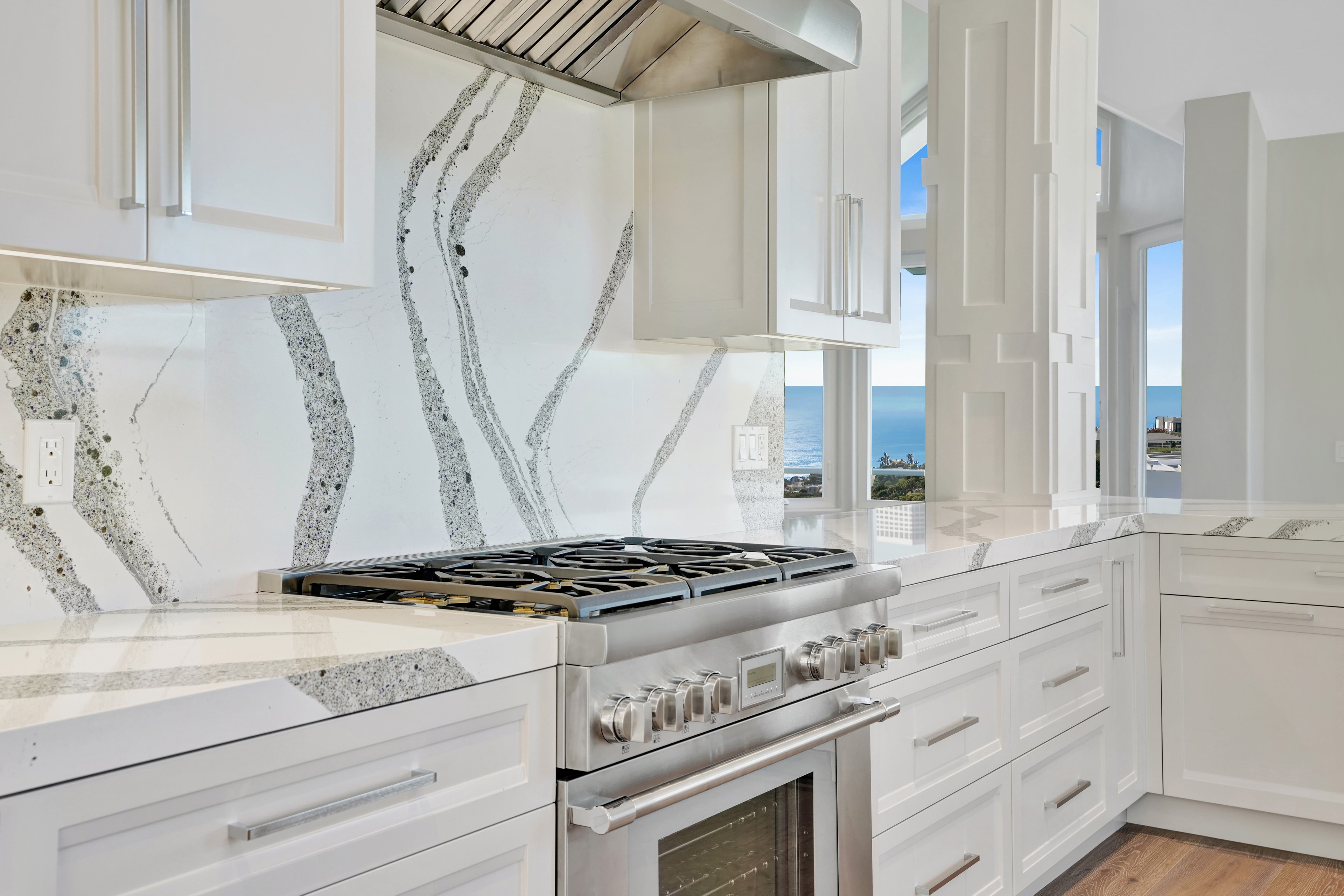 Kitchen remodel in Laguna Beach CA — closeup of white farmhouse apron sink with chrome gooseneck pull-down faucet, book-matched quartz slab backsplash, and panoramic ocean view through casement window by Main Street Kitchen & Flooring